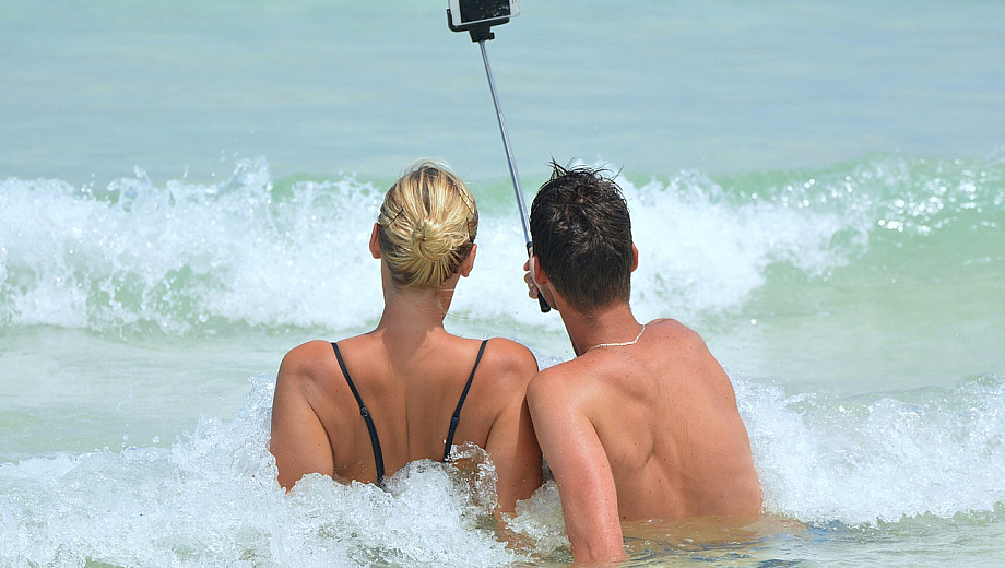 Zwei Menschen sitzen am Strand in den Wellen und machen ein Selfie von sich.