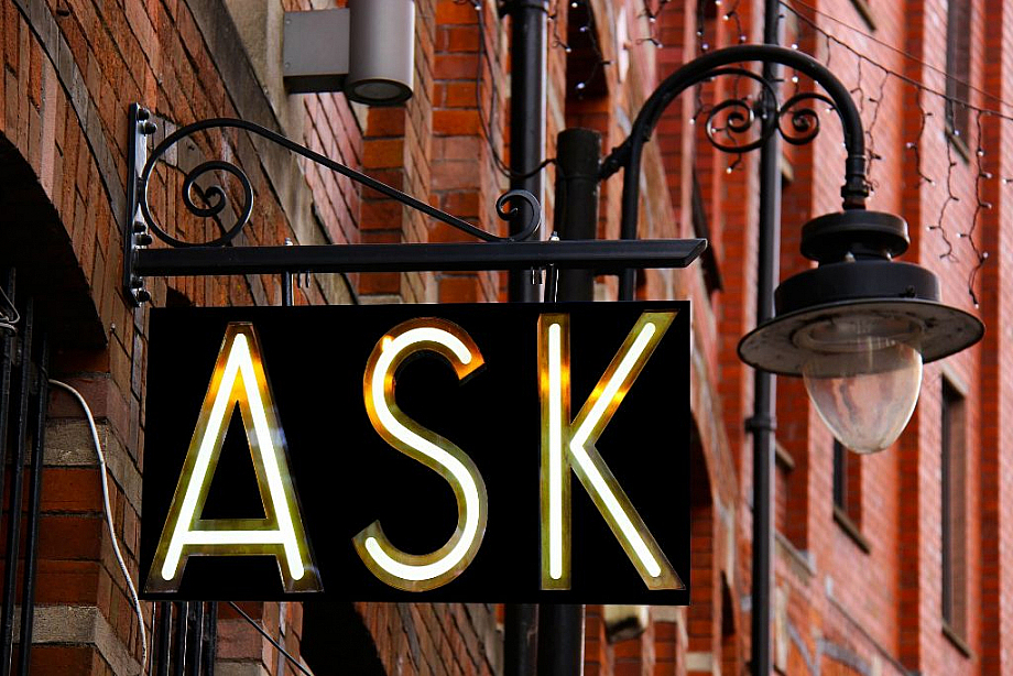 In großen Buchstaben steht das Wort "Ask" (englisch für fragen) auf einem Schild neben einer Laterne.