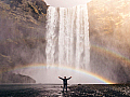 Eine Person steht unter einem Wasserfall. Im Sprühnebel leuchtet ein Regenbogen.