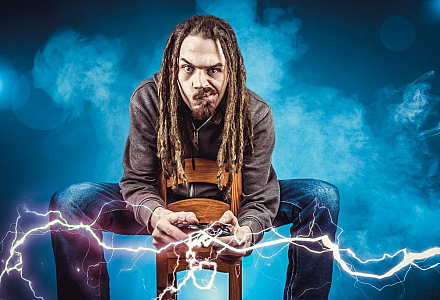 Ein Mann mit Dreadlocks sitzt auf einem Hocker, beugt sich nach vorne und hält einen Spiele-Controller in der Hand, aus dem Blitze schließen. Der Gamer guckt hoch konzentriert.