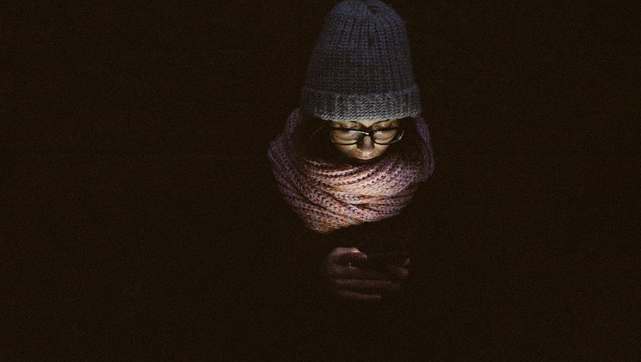 Eine Frau mit Mütze und Schal guckt in ihr Smartphone, während um sie herum alles schwarz ist.