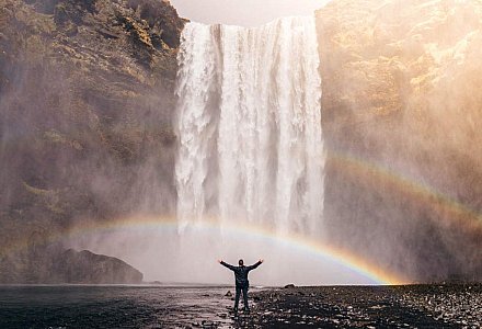 Eine Person steht unter einem Wasserfall. Im Sprühnebel leuchtet ein Regenbogen.