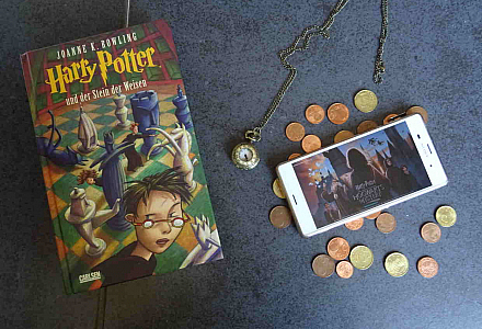 Auf einem Tisch liegt ein Harry Potter Roman, daneben eine Uhr, ein bisschen Kleingeld und ein Smartphone mit dem Mobile Game Hogwarts Mystery.
