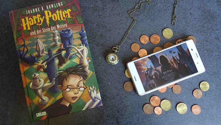 Auf einem Tisch liegt ein Harry Potter Roman, daneben eine Uhr, ein bisschen Kleingeld und ein Smartphone mit dem Mobile Game Hogwarts Mystery.