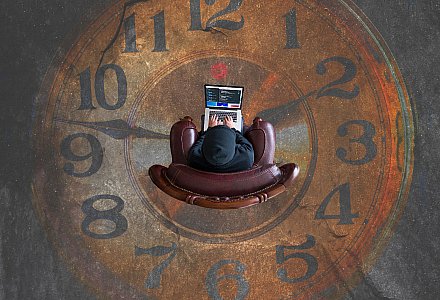Mensch sitzt vor einem Laptop, im Hintergrund ist einen große Uhr zu sehen.