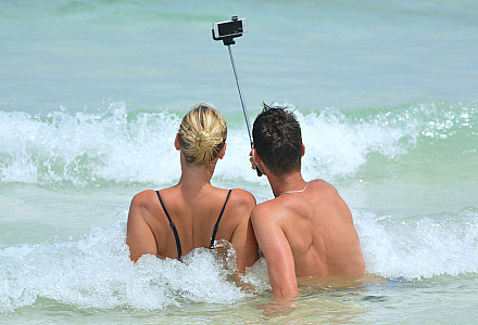 Zwei Menschen sitzen am Strand in den Wellen und machen ein Selfie von sich.