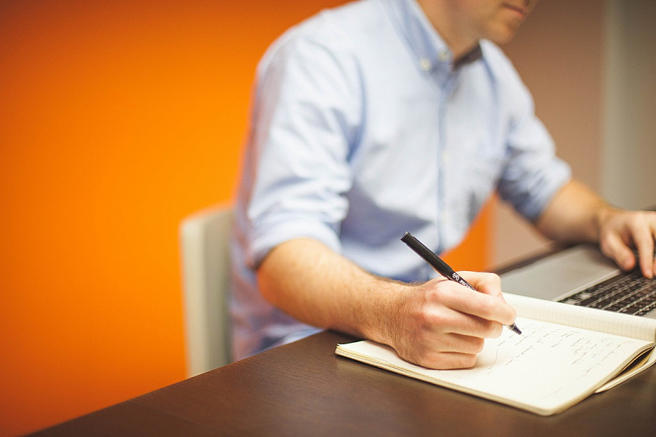 Ein Mann sitzt am Schreibtisch, eine Hand auf der Tastatur, in der anderen Hand einen Stift. Er schreibt etwas in ein Notizheft.