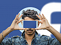 Person hält ein Smartphone vor sein Gesicht. Der Bildschirm leuchtet blau. Im Hintergrund erkennt man das Facebook-Logo.