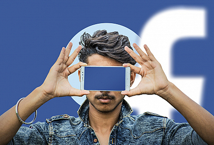 Person hält ein Smartphone vor sein Gesicht. Der Bildschirm leuchtet blau. Im Hintergrund erkennt man das Facebook-Logo.