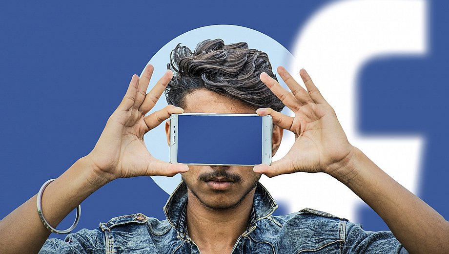 Person hält ein Smartphone vor sein Gesicht. Der Bildschirm leuchtet blau. Im Hintergrund erkennt man das Facebook-Logo.