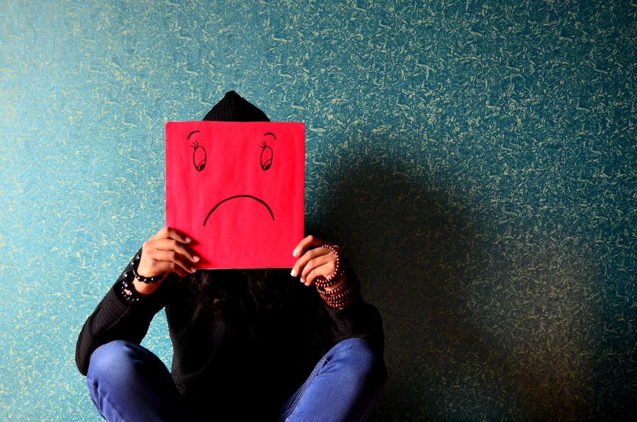 Person hockt auf dem Boden an die Wand gelehnt und hält sich ein rotes Schild vors Gesicht, auf dem ein trauriger Smiley zu sehen ist.