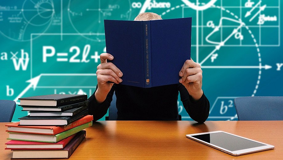 Dozent hinter einem Tisch voll Bücher. Im Hintergrund eine Tafel mit vielen Formeln.
