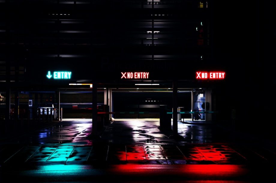 Einfahrt zu einem Parkhaus. Es ist sehr dunkel. Im Foto stehen die leuchtenden Schilder über den drei Durchfahrten hervor. Die linke Durchfahrt ist auf grün gestellt und somit als Einfahrt gekennzeichnet. Die mittlere und rechte Druchfahrt sind rot leuchtende Ausfahrten.