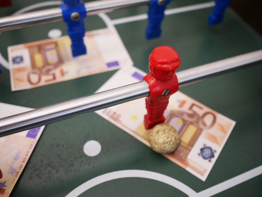 Tischkicker in der Nahaufnahme. Man sieht eine rote SPielfigur mit einem goldenen Ball vor den Füßen. Auf dem Spielfeld liegen 50-Euro-Scheine. 