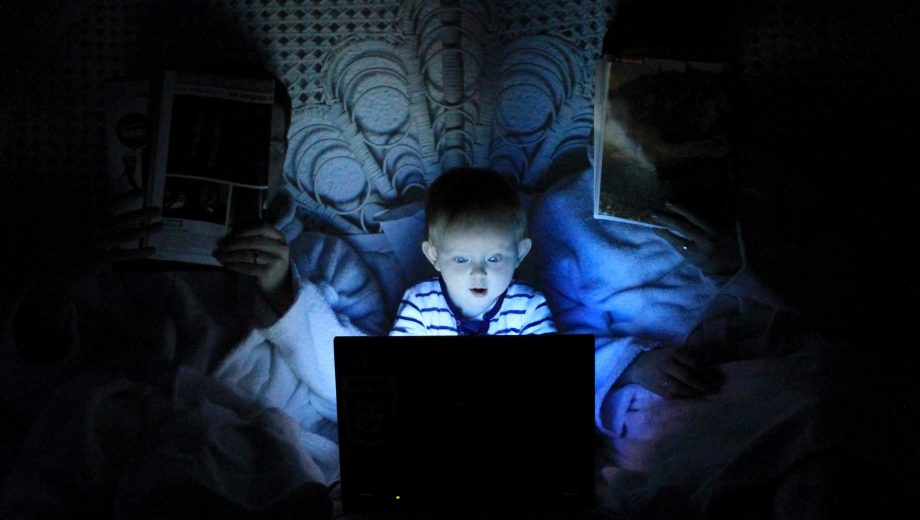 Kind liegt mit zwei Erwachsenen im Bett. Die Erwachsenen lesen Zeitung. Das Kinf guckt auf einen hell erleuchteten Bildschirm.