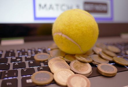 Auf einer Tastatur liegen ein Tennisball und viele Euro-Mümzen. Im Hintergrund zeigt der Bildschirm das Wort 