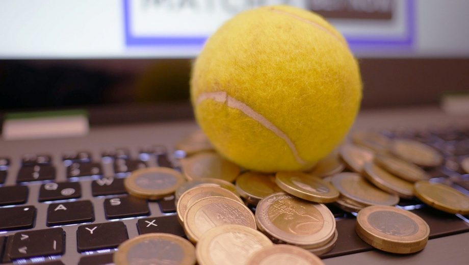 Auf einer Tastatur liegen ein Tennisball und viele Euro-Mümzen. Im Hintergrund zeigt der Bildschirm das Wort "Match".