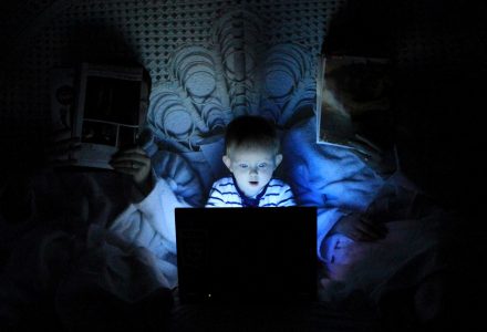 Kind liegt mit zwei Erwachsenen im Bett. Die Erwachsenen lesen Zeitung. Das Kinf guckt auf einen hell erleuchteten Bildschirm.
