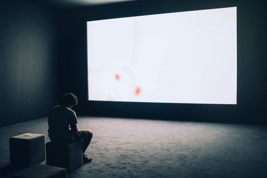 Person sitzt alleine in einem großen Raum mit riesigem Bildschirm an der Wand