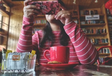 Frau am, Tisch, vor ihr ein Kaffee, hält das Smartphone vors Gesicht